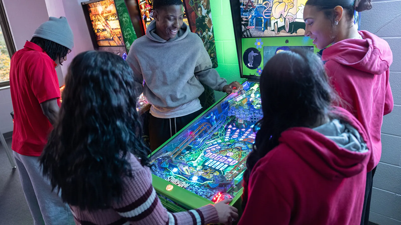 A group of people stands around a green pinball machine in an arcade.