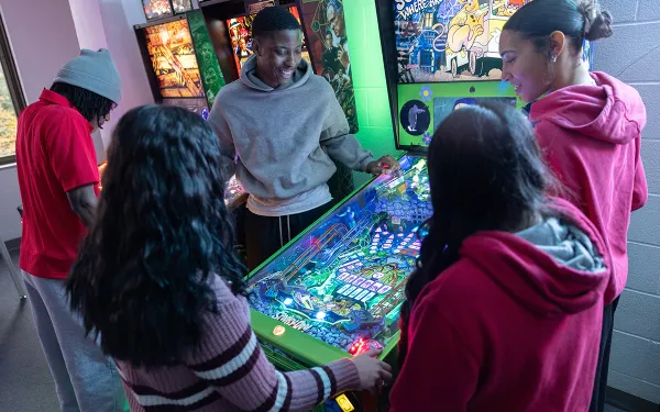 A group of people stands around a green pinball machine in an arcade.