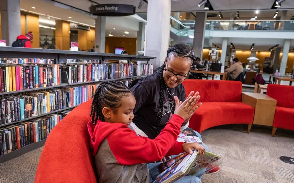 A mother and child read books in Thompson library, surrounded by shelves of books