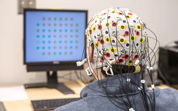 A person wearing a white cap with multicolored circles and wires looks at a computer monitor displaying multicolored dots.